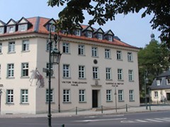 Ein helles dreistöckiges Gebäude mit einem roten Dach in dem viele Erker eingebaut sind, wird als Geschäftsstelle genutzt. 